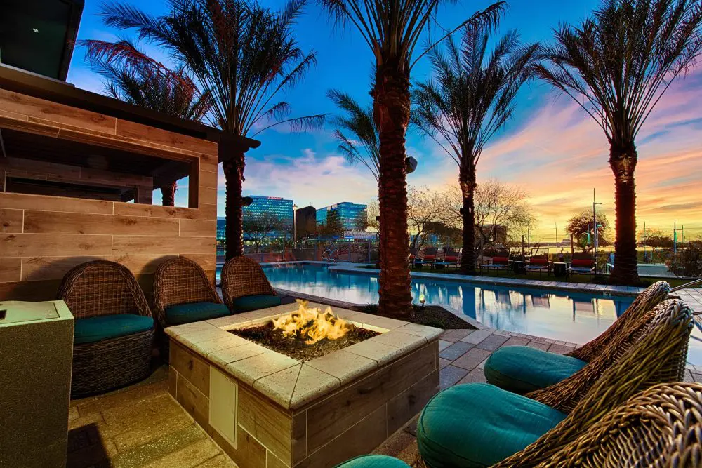 Tempe, AZ Luxury Apartments - Vela At Tempe Town Lake - Poolside Lounge Area With Raised Firepit, Outdoor Seating, Palm Trees, And City View